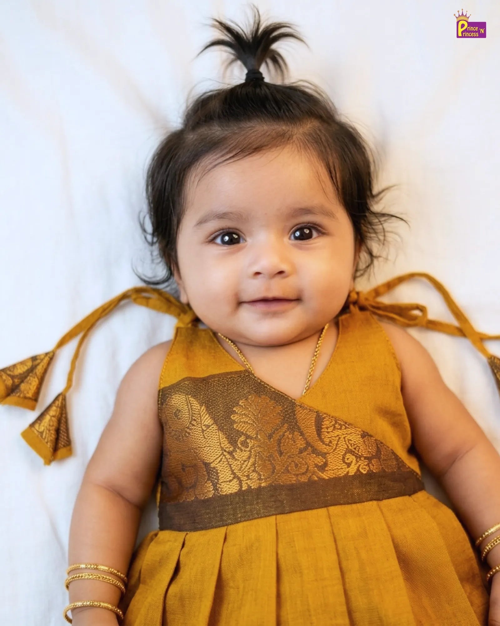 Baby wearing a mustard yellow dress with intricate designs on a white background