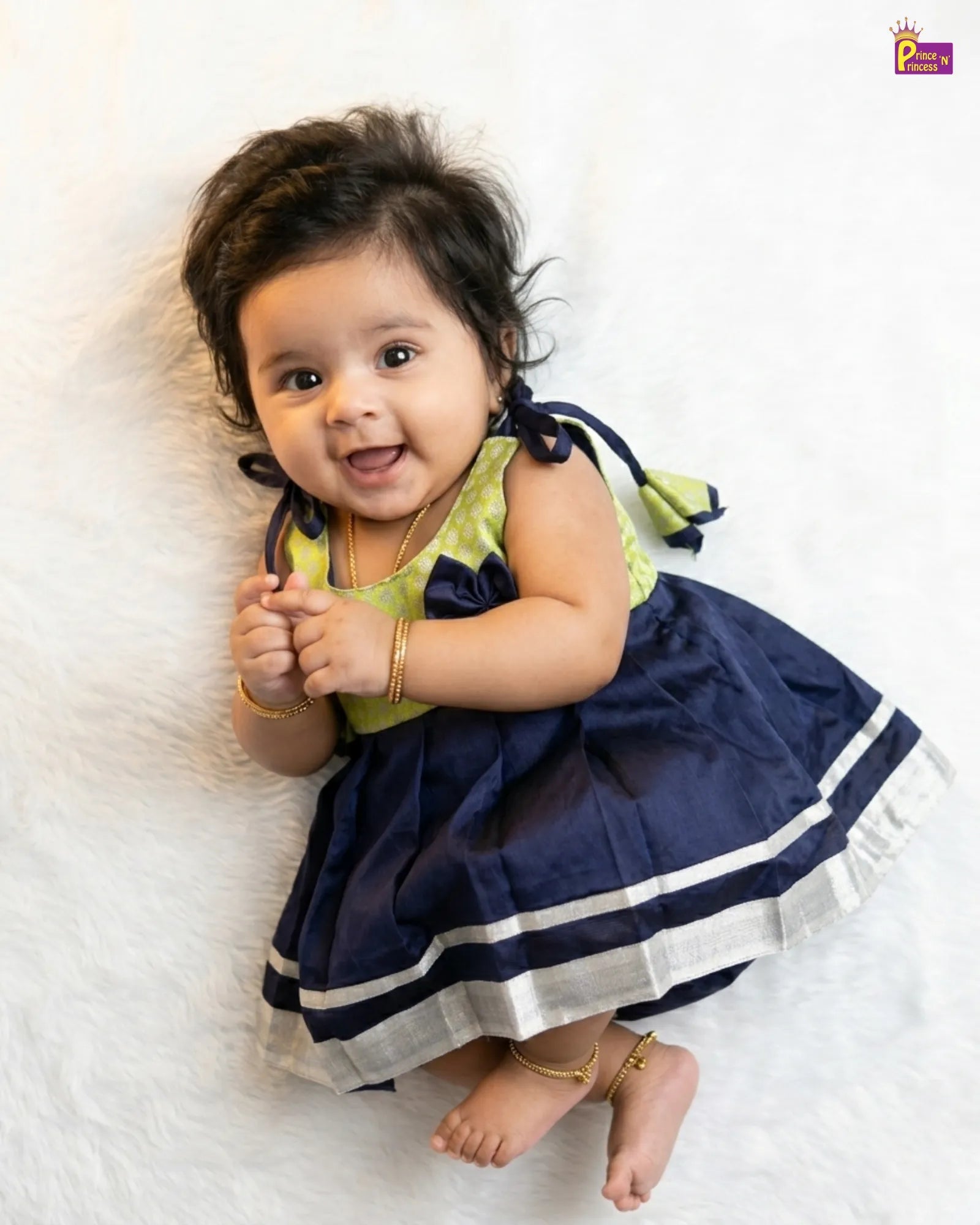 Baby in a green and navy dress sitting on a white surface