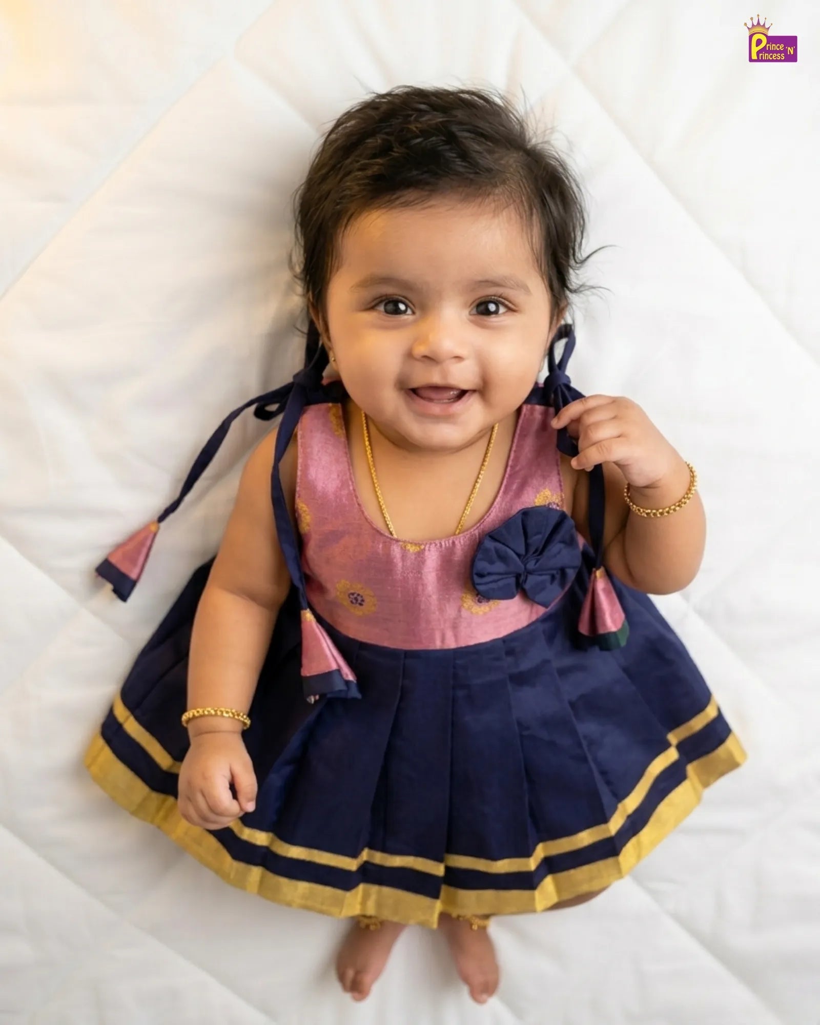 Baby wearing a pink and navy dress with a bow, lying on a white surface.