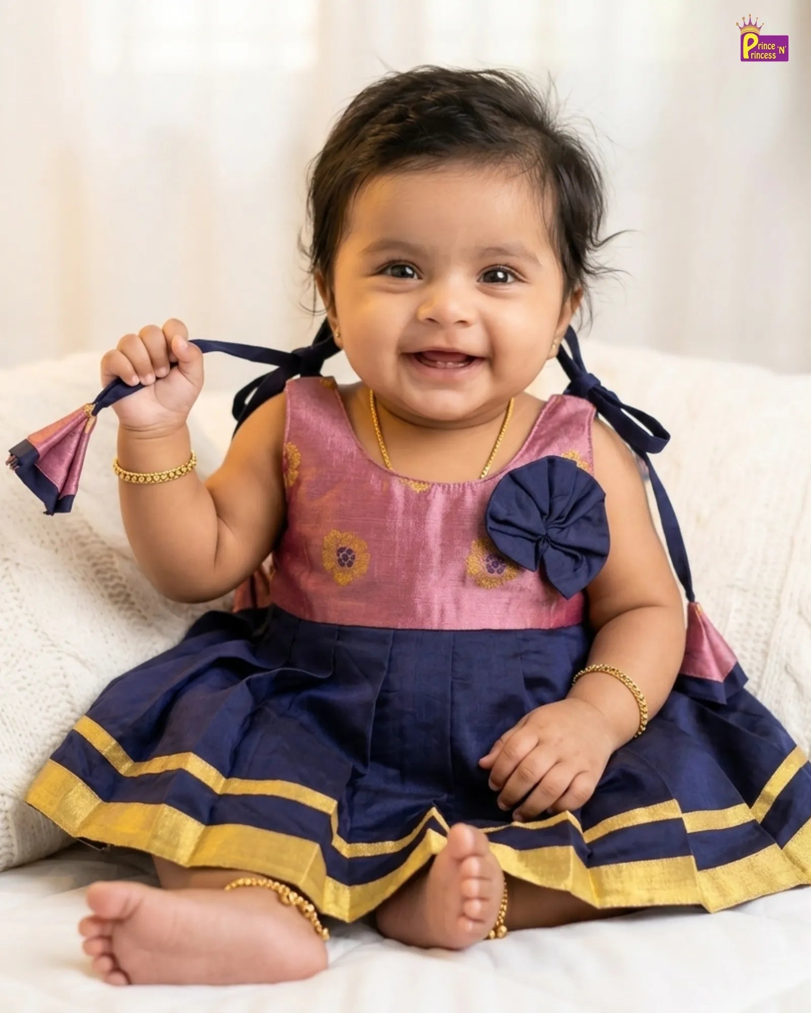 Baby in a pink and navy dress with a flower detail, sitting on a white surface.