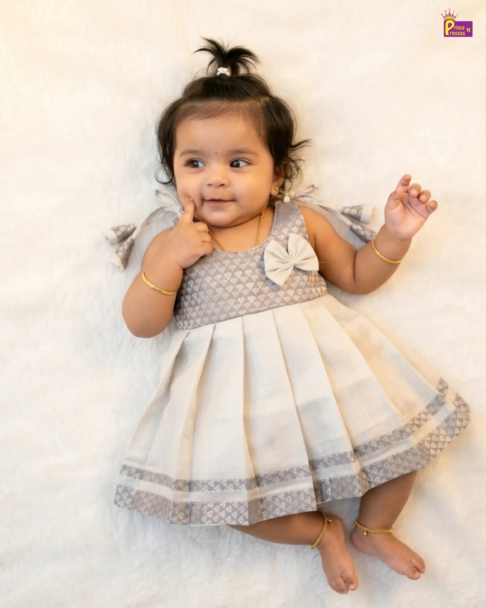 Baby in a gray and white dress with a bow, lying on a white surface.