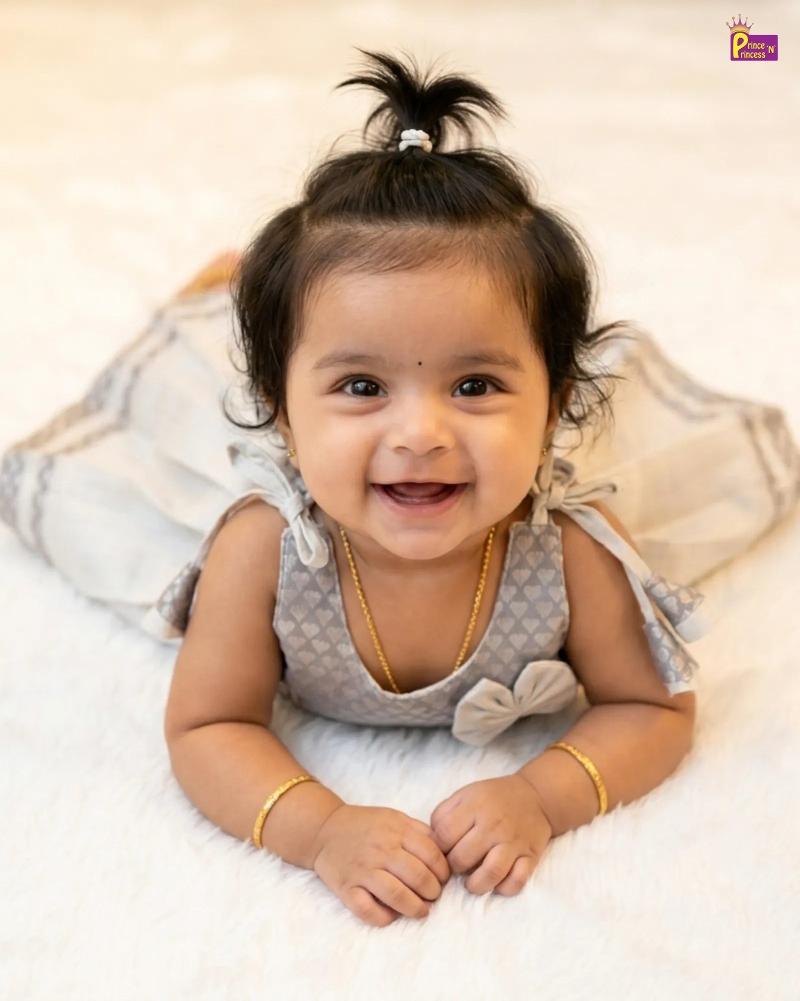 Smiling baby girl wearing a gray dress with ruffles on a white blanket