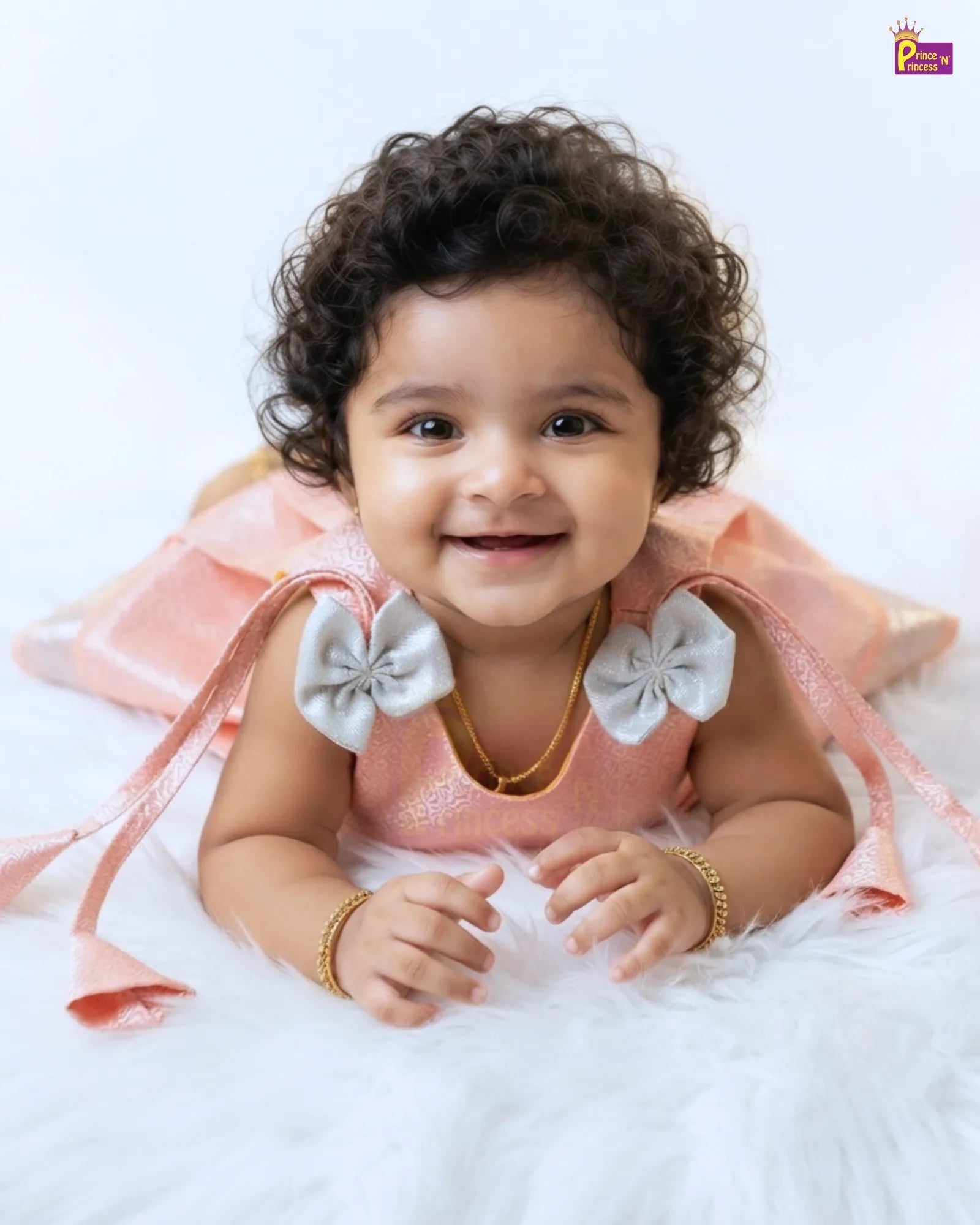 A smiling baby girl with curly dark hair lies on a white fluffy rug wearing a light pink dress with large white bow details on the shoulders and gold jewelry.