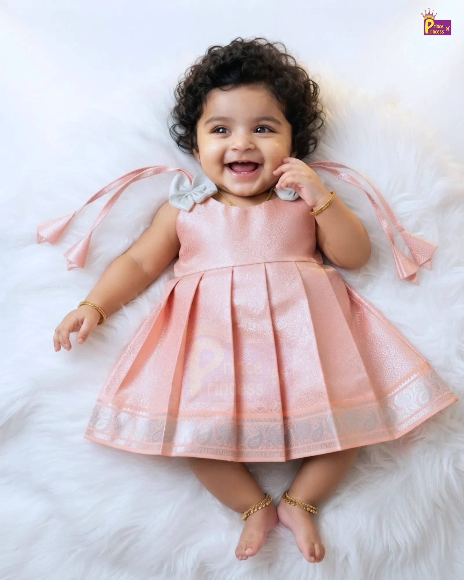 A smiling toddler with curly dark hair lies on a white faux-fur surface wearing a sleeveless pleated light pink dress with ribbon ties at the shoulders and a decorative border at the hem.