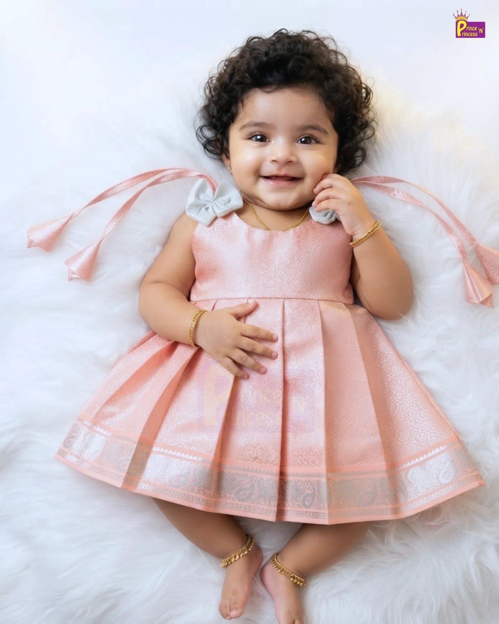 A smiling toddler with curly dark hair lies on a white faux-fur surface wearing a sleeveless peach-pink traditional dress with a pleated skirt, a light gray bow at the neckline, and long ribbon tails. The child is also wearing gold jewelry.