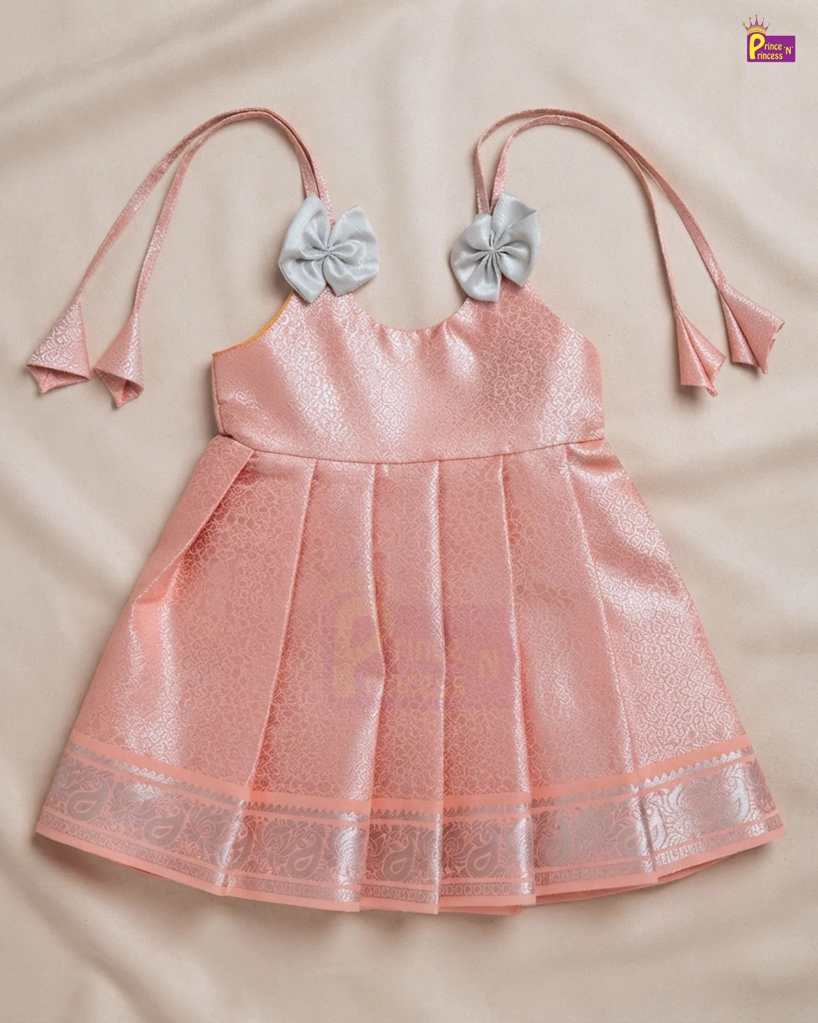 A pink pleated baby dress with a shimmering texture, featuring a decorative silver-gray bow on each strap and a matching pink bow tie at the neckline, laid flat on a beige fabric background.