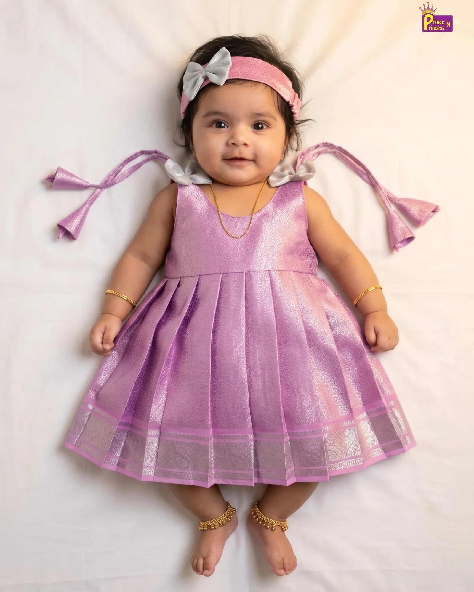 A baby girl lying on a white sheet wearing a shiny lavender-purple sleeveless dress with a pleated skirt, a pink headband with a white bow, and gold jewelry.