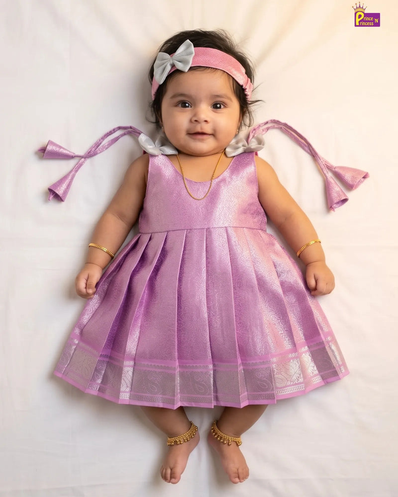A baby girl lying on a white sheet wearing a shiny lavender-purple sleeveless dress with a pleated skirt, a pink headband with a white bow, and gold jewelry.