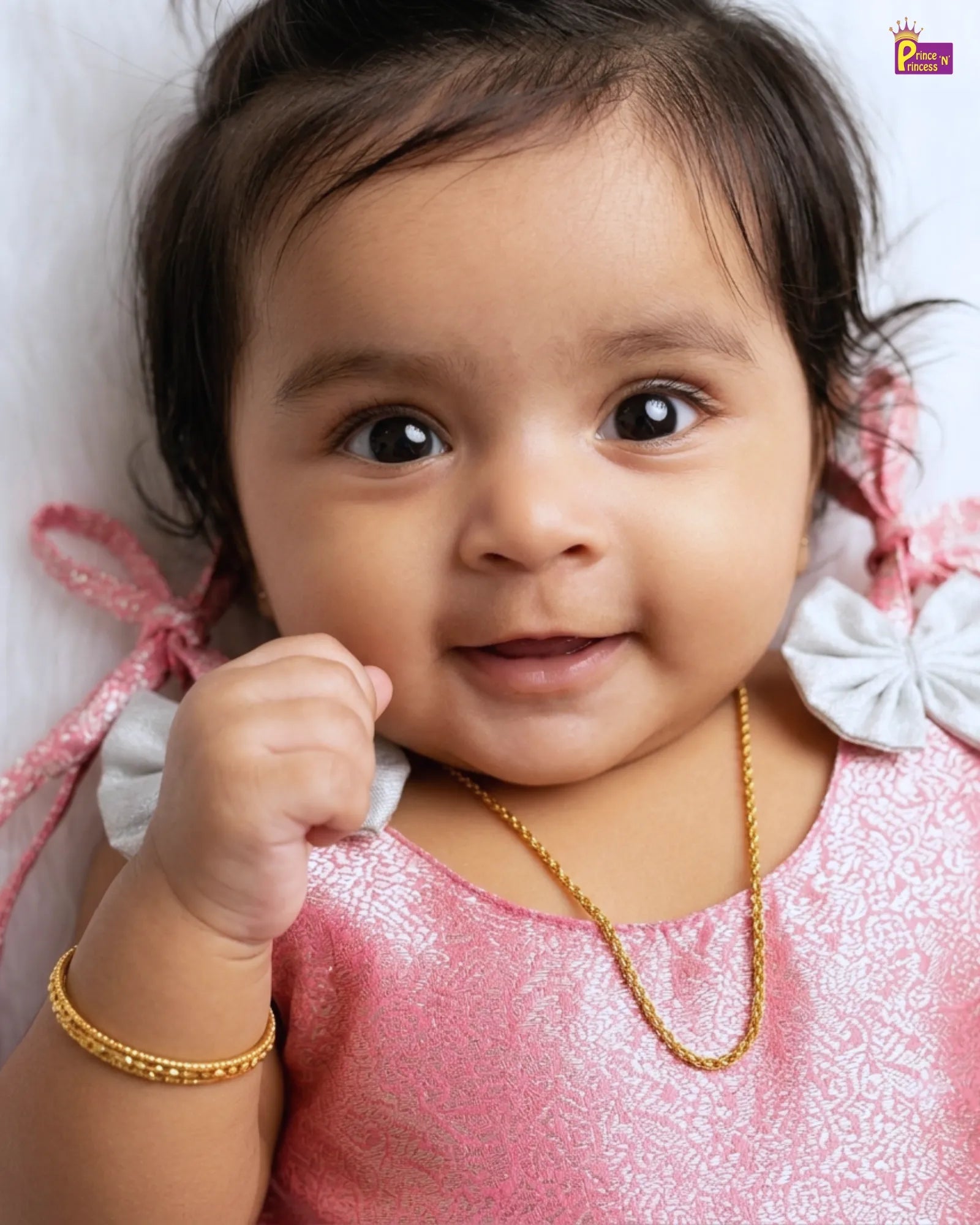 Baby in a pink outfit with a gold necklace and bracelet, lying on a white background.