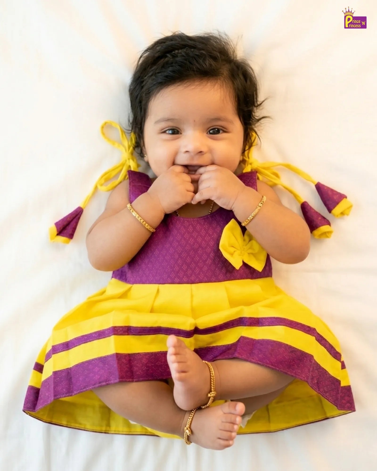 Baby wearing a yellow and purple dress lying on a white surface