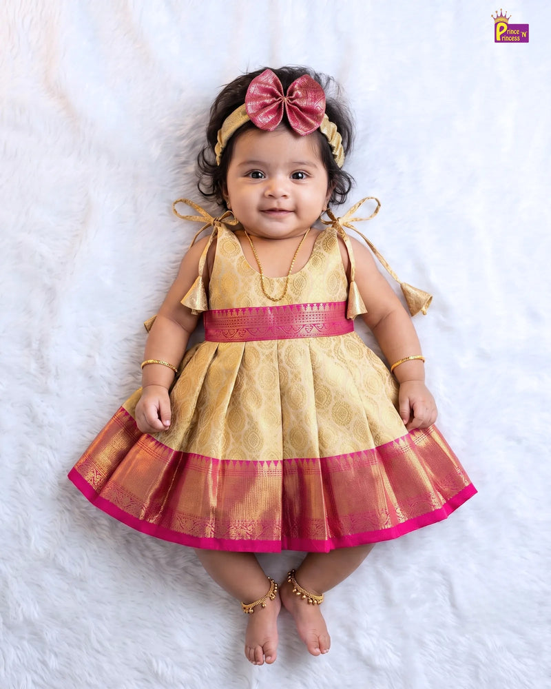 A baby girl wearing a traditional Indian gold and pink dress with a matching bow headband, lying on a white faux fur background.