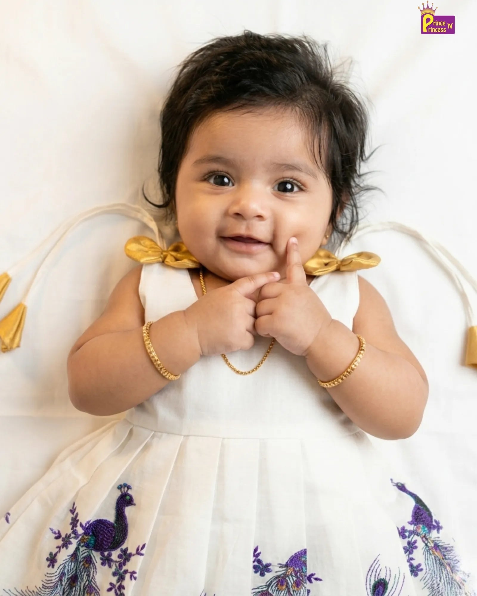 Baby wearing a white dress with peacock patterns and gold accents on a white background