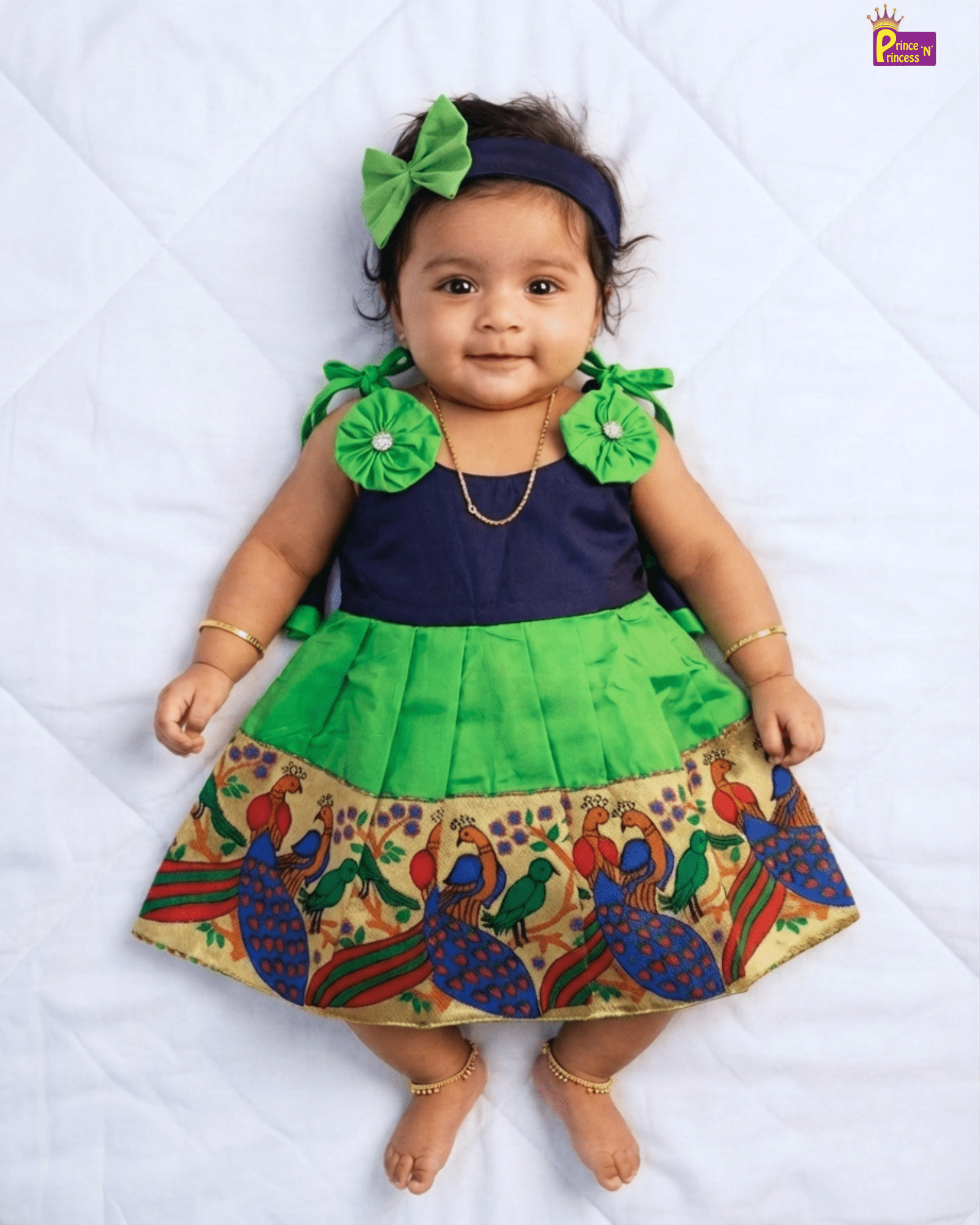 A baby girl wearing a traditional Indian dress with a navy blue top, a bright green pleated skirt, and a peacock patterned border, lying on a white quilted blanket. She is also wearing a green bow headband, gold bangles, and a gold necklace.