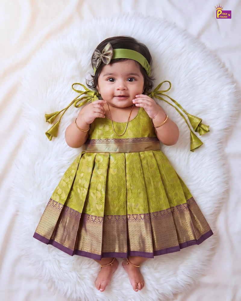A baby girl wearing a green and gold traditional Indian dress with a matching headband, lying on a white faux fur rug.