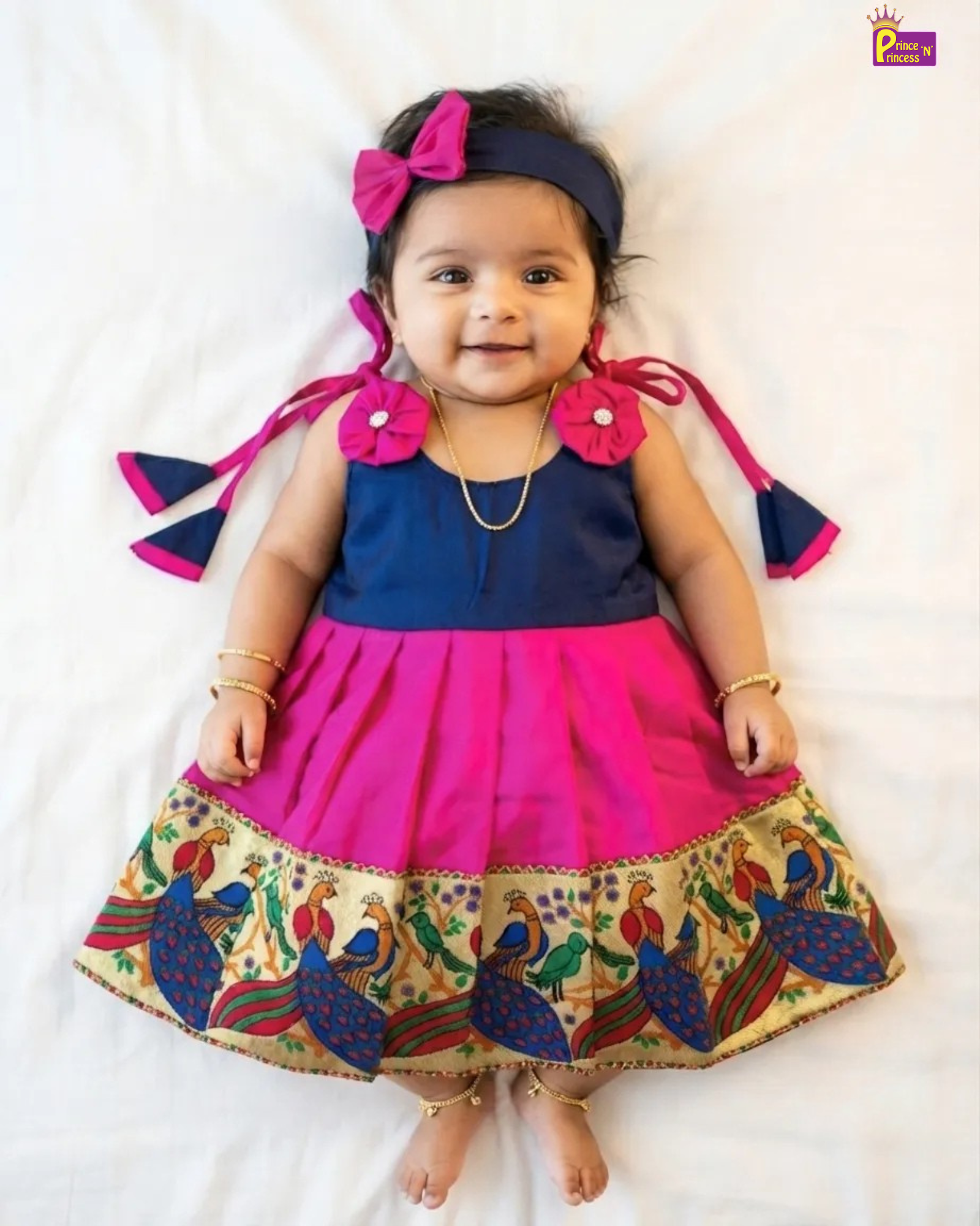 A toddler girl lying on a white sheet wearing a two-tone navy blue and hot pink dress with a peacock patterned hem, a matching pink bow headband, and gold jewelry.