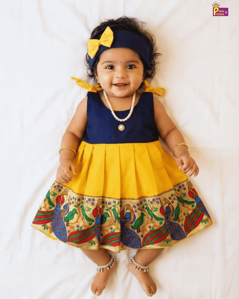 A baby girl wearing a yellow and navy blue dress with a colorful peacock pattern on the hem, a matching yellow bow headband, and a pearl necklace, lying on a white sheet.