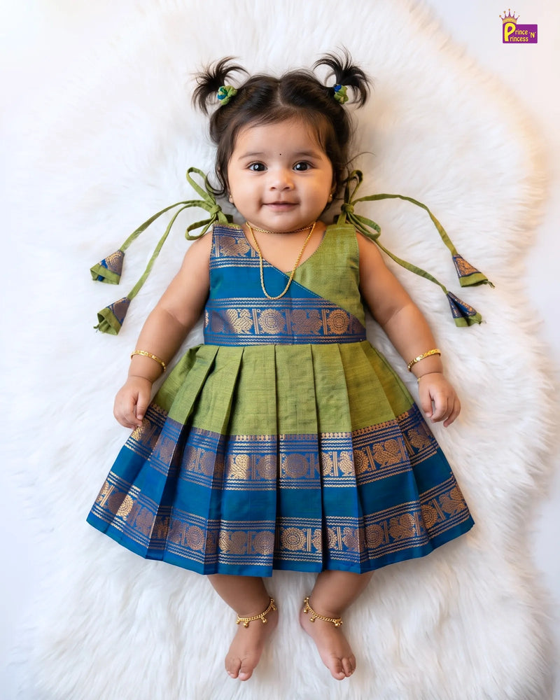 A toddler girl wearing a sleeveless pleated dress with green and blue borders, lying on a white faux fur rug. The dress has a green upper bodice, a blue waistband with gold zari detailing, and a blue pleated skirt with matching gold borders.