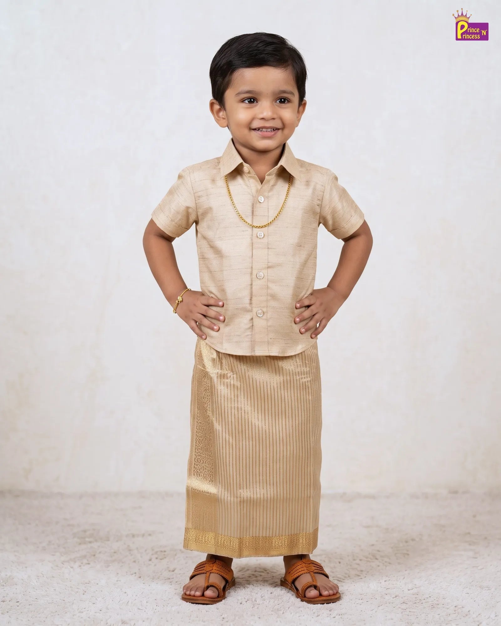A young boy modeling a traditional Indian outfit consisting of a short-sleeve beige kurta and a matching pleated kurta skirt, accessorized with a gold chain necklace and brown leather sandals, set against a textured off-white background.
