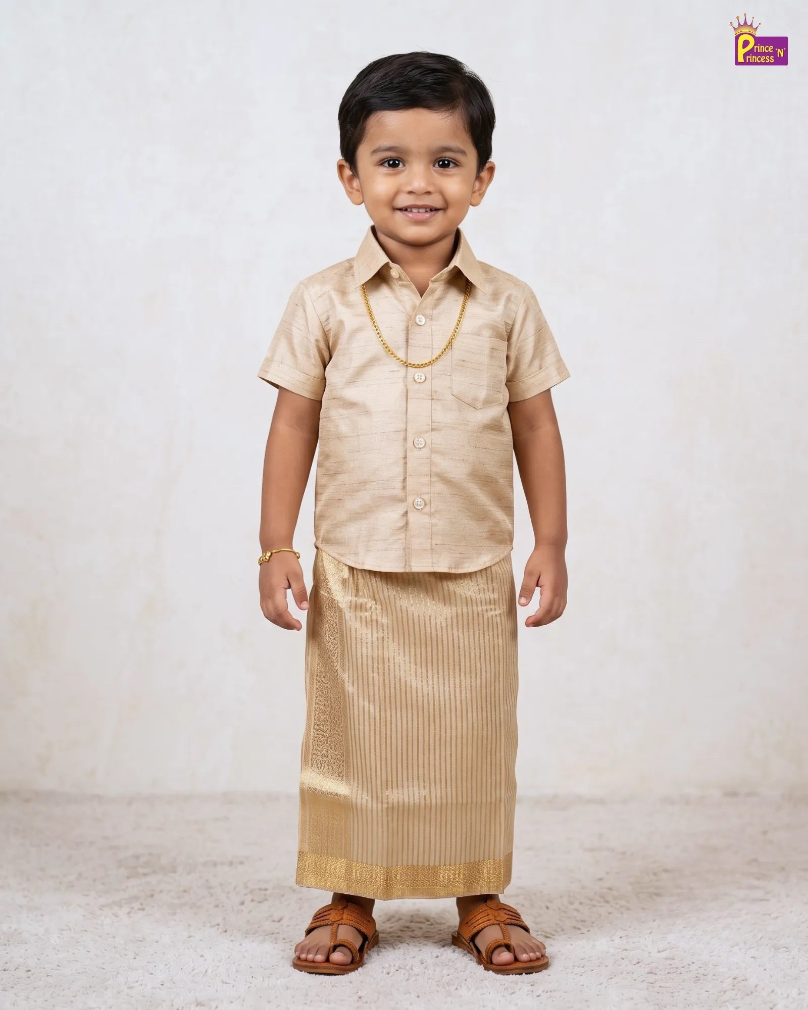 A young boy modeling a traditional Indian outfit consisting of a light beige short-sleeve button-up shirt and a matching vertically striped kurta, styled with a gold necklace and brown leather sandals against a textured off-white background.