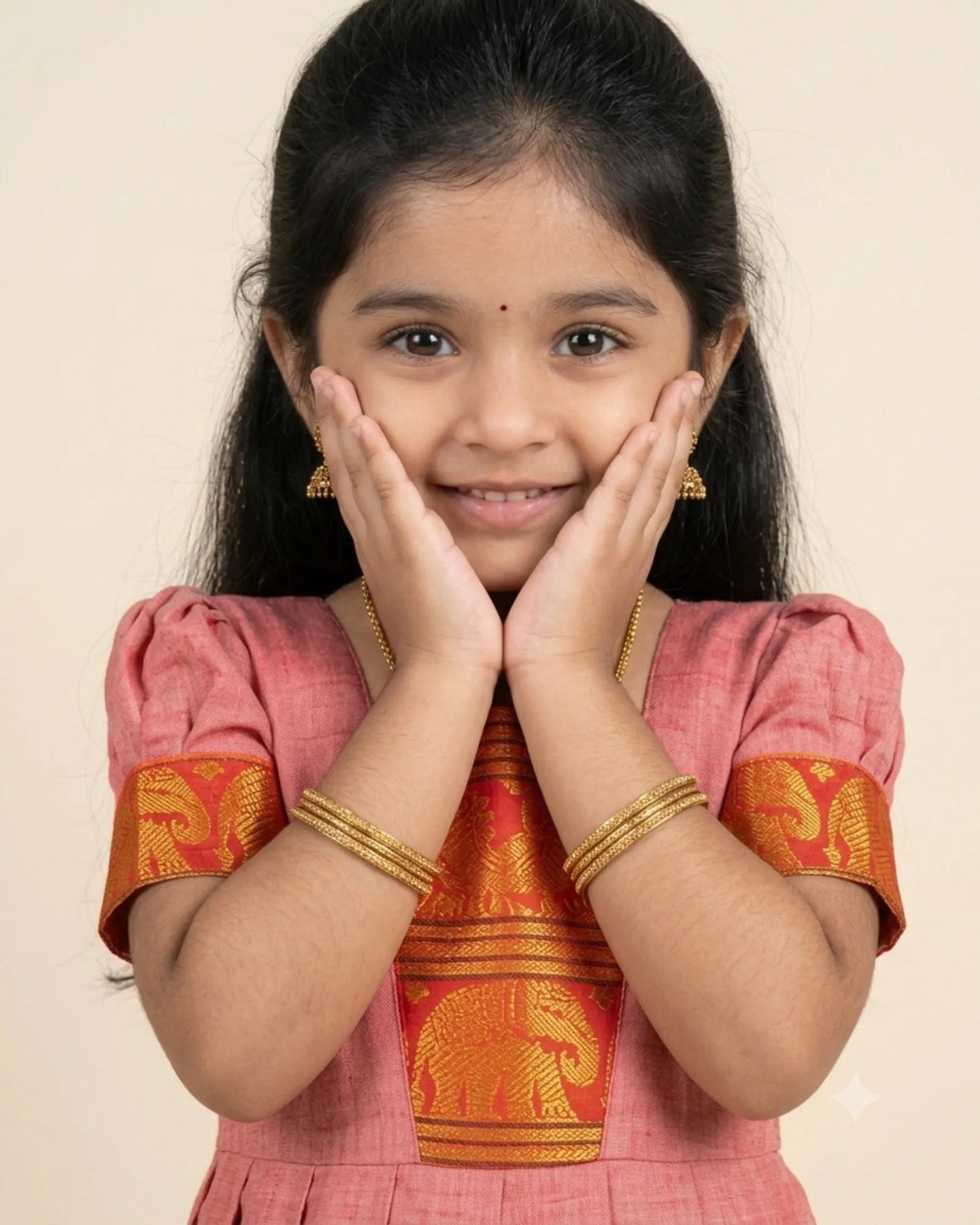 A young girl with dark hair wearing a pink and orange traditional Indian dress with gold embroidery, posing with her hands on her cheeks against a plain beige background.