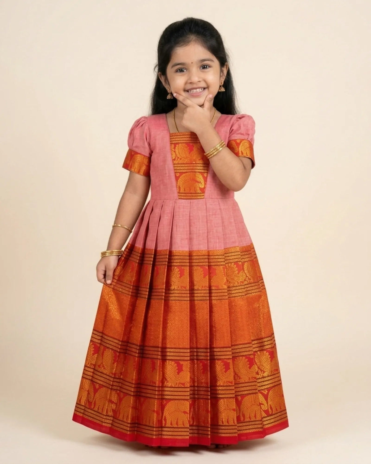 A young girl modeling a traditional Indian salwar kameez dress with a pink upper bodice and an orange lower skirt featuring woven elephant patterns, set against a plain beige background.