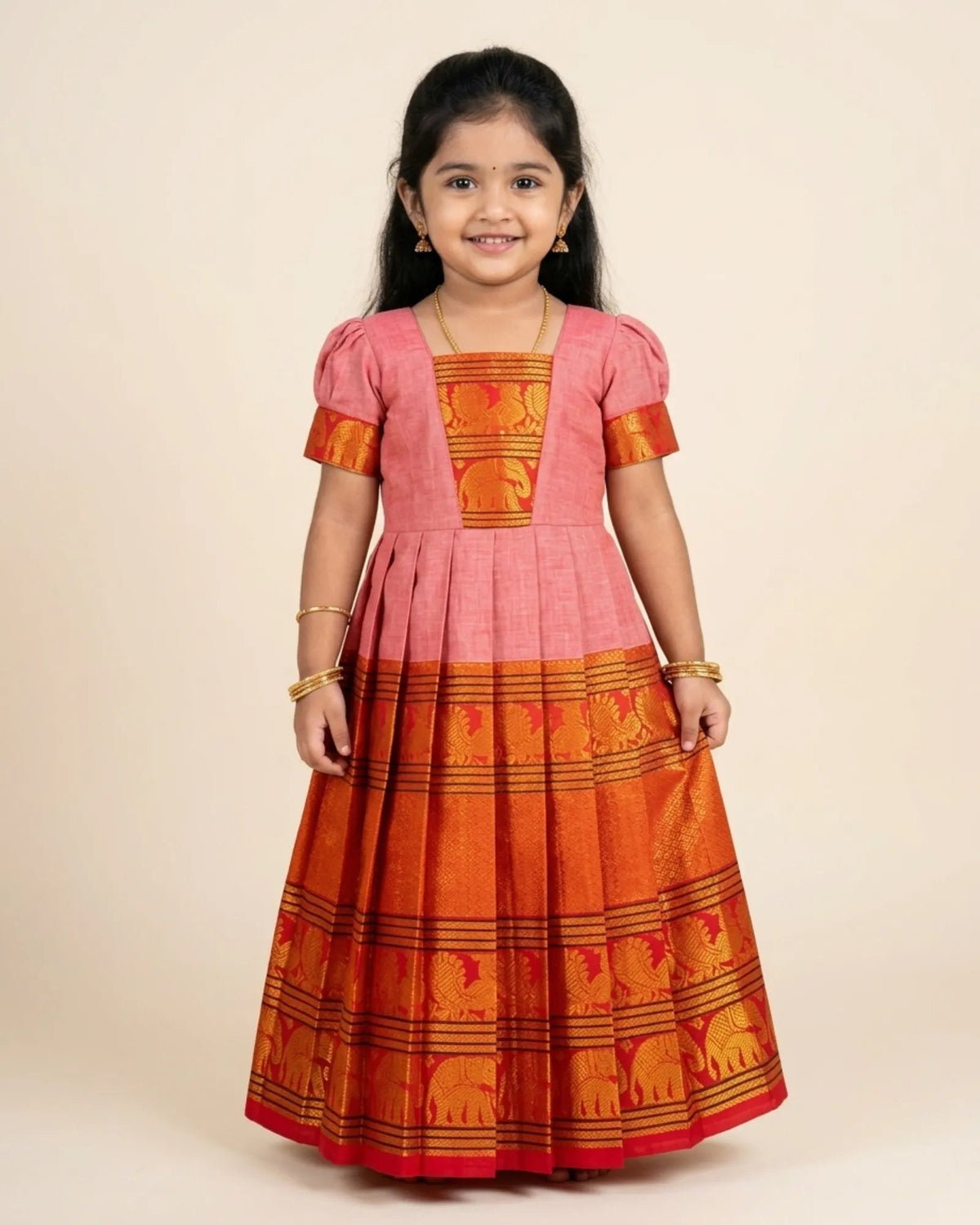 A young girl modeling a traditional Indian salwar kameez dress with a pink upper bodice and a wide orange and red lower skirt featuring elephant motifs.