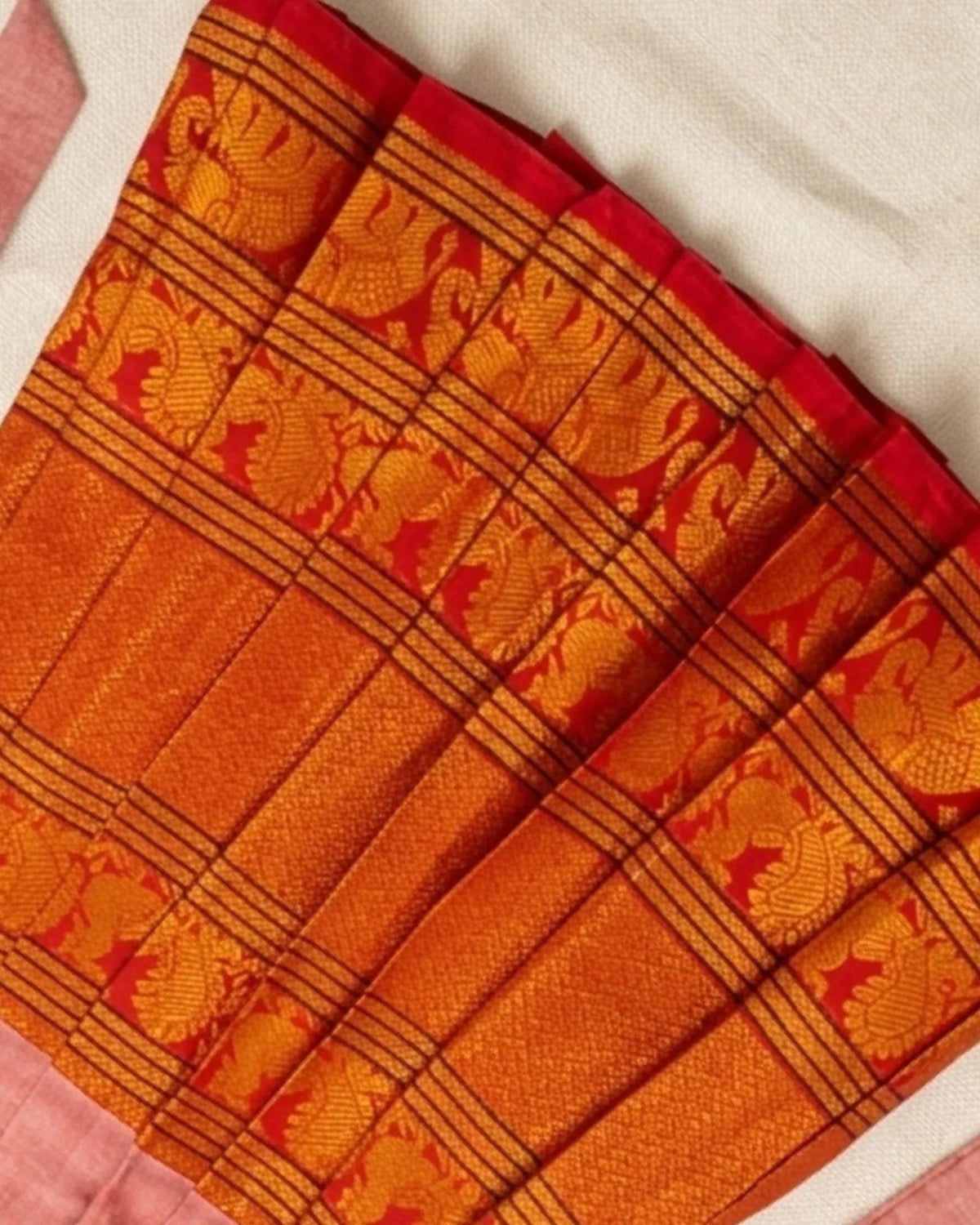 A close-up of a folded orange and red patterned fabric, likely a saree or textile, showing woven floral motifs and striped borders on a light neutral background.