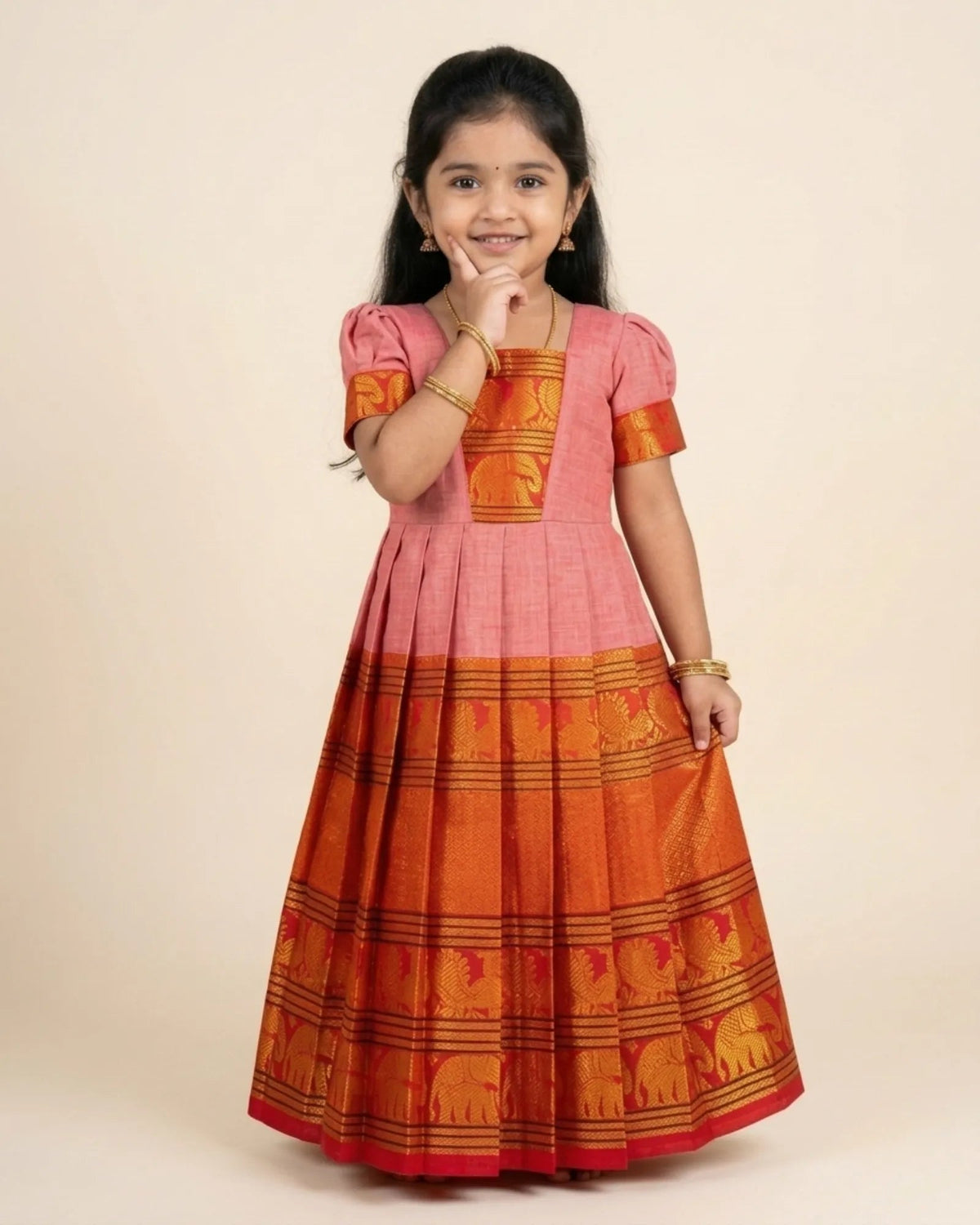 A young girl modeling a traditional Indian salwar kameez dress with a pink upper bodice and a wide orange skirt featuring woven elephant motifs, set against a plain beige background.