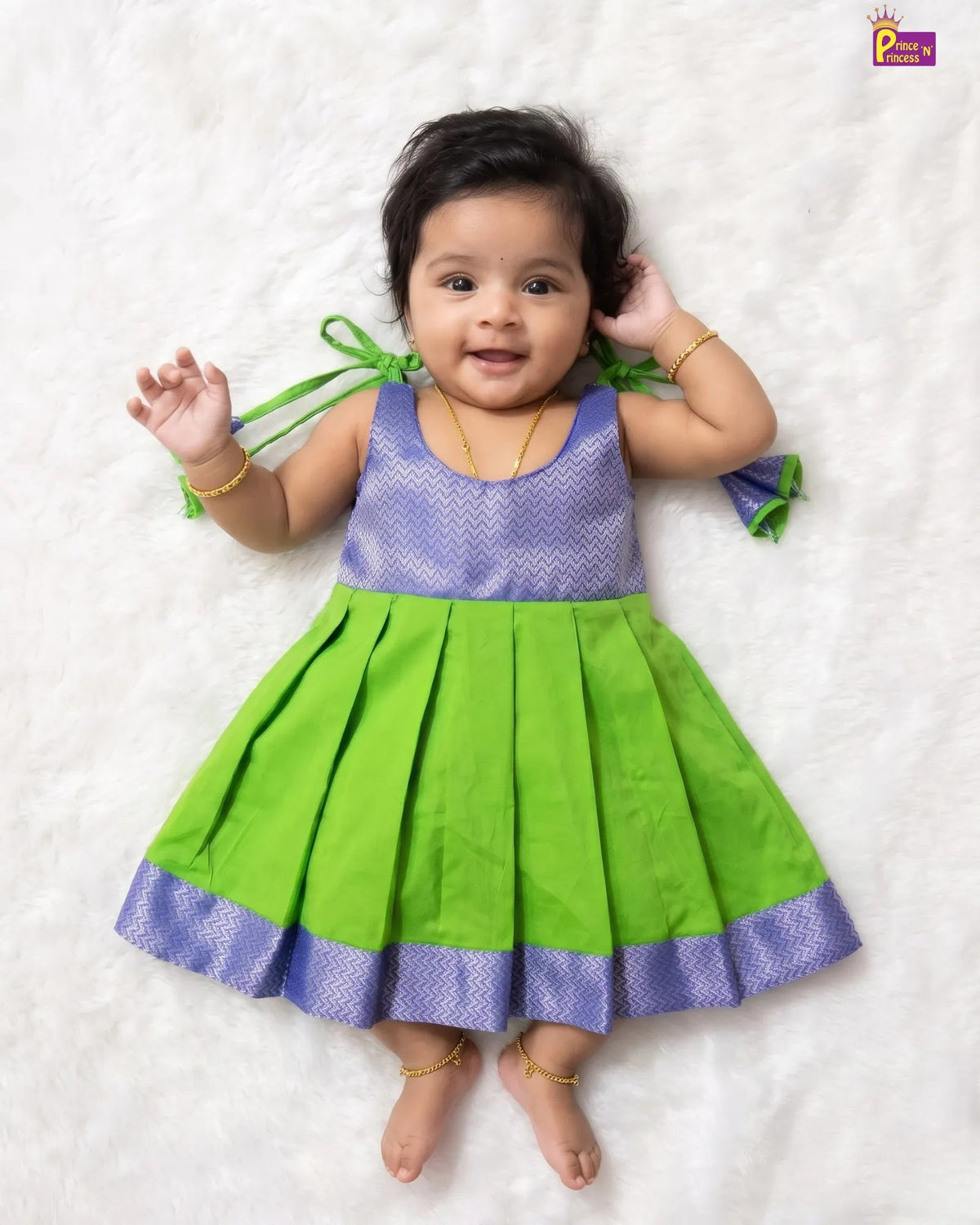 A baby girl lying on a white textured blanket, wearing a green and purple traditional Indian dress with a pleated skirt and patterned purple bodice.
