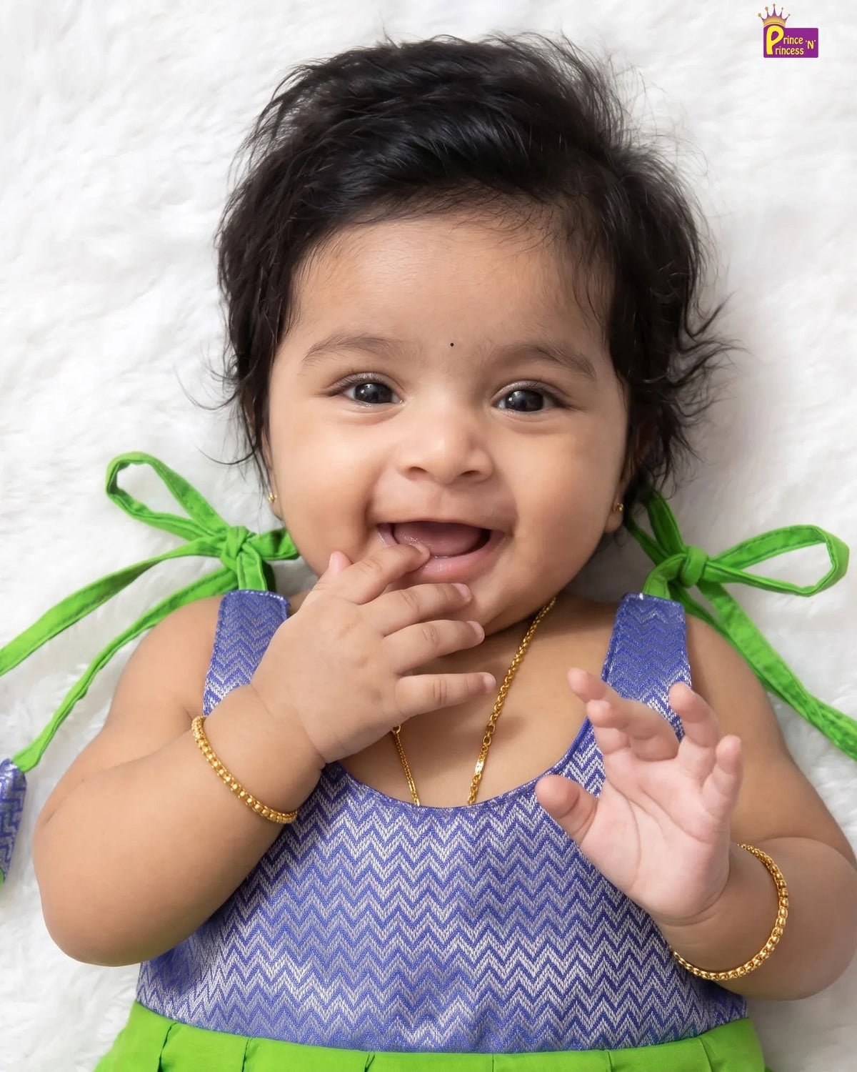 A baby lying on a white textured blanket, wearing a purple and white chevron-patterned top with bright green ties and a matching green bottom, accessorized with gold bracelets and a gold necklace.