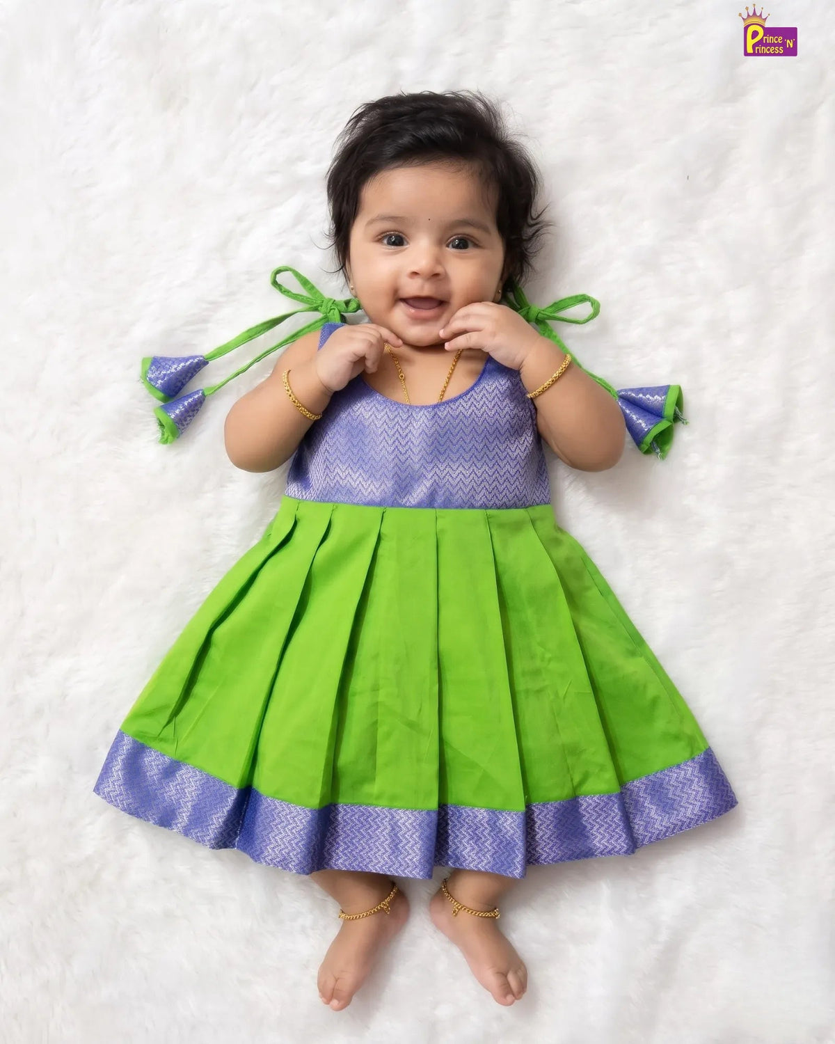 A baby girl wearing a bright green and purple traditional dress, lying on a white textured blanket. The dress has a purple patterned bodice, green pleated skirt, and matching green ribbon ties on the sides.