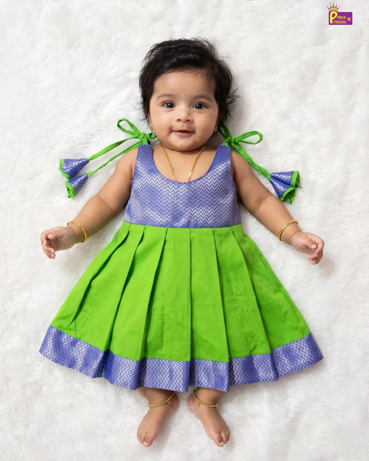 A baby girl lying on a white textured blanket, wearing a sleeveless green and purple dress with pleated skirt and bow details on the shoulders.