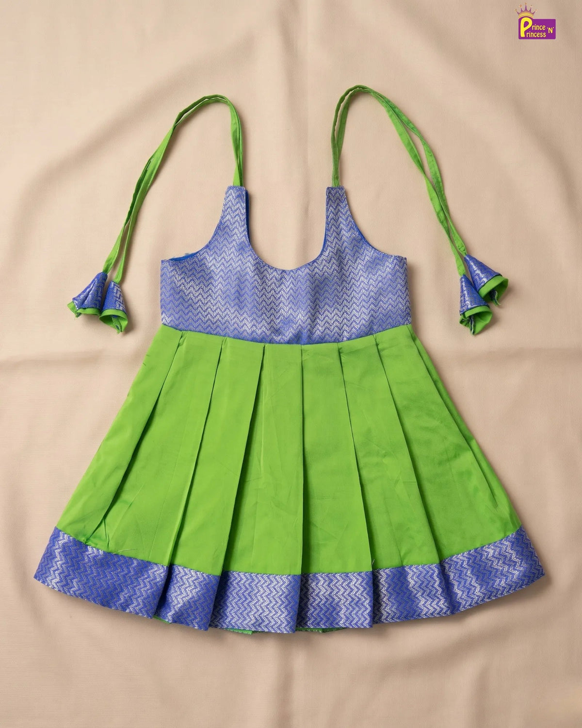 A sleeveless children's dress with a bright green pleated skirt and a blue patterned bodice, featuring green tie straps, laid flat on a beige fabric background.