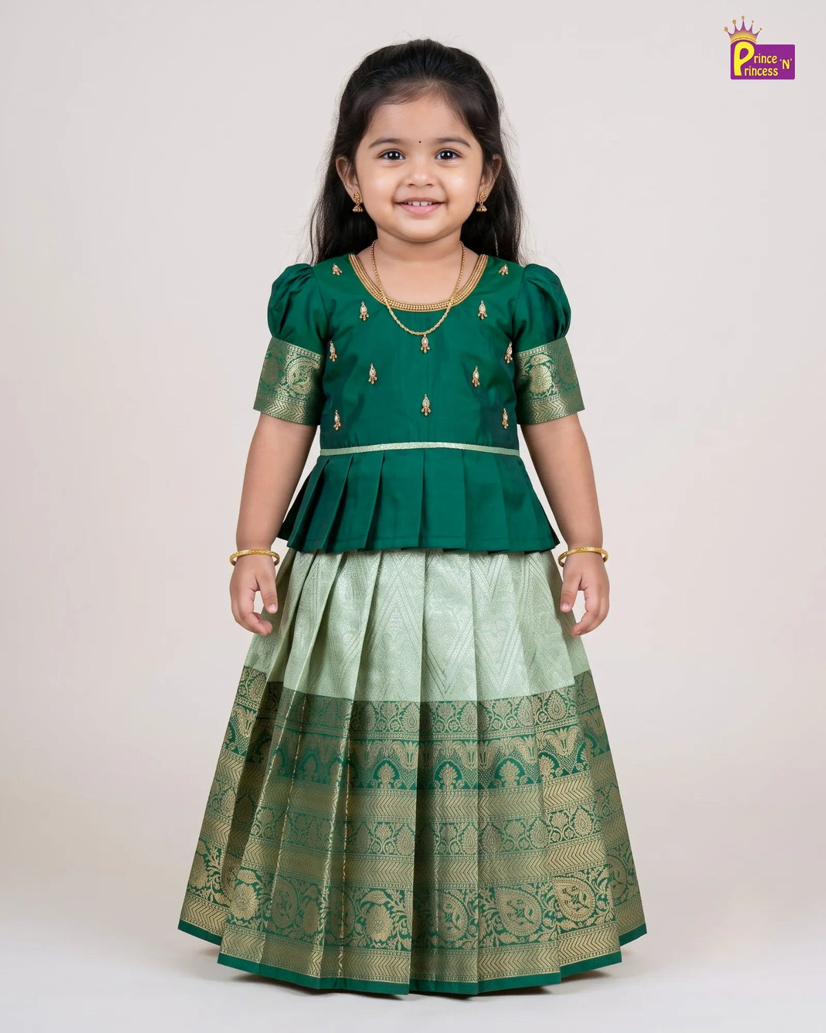 A young girl modeling a traditional Indian ethnic outfit consisting of a dark green peplum top with gold embroidery and a light green pleated skirt with a wide gold border, set against a plain light beige background.