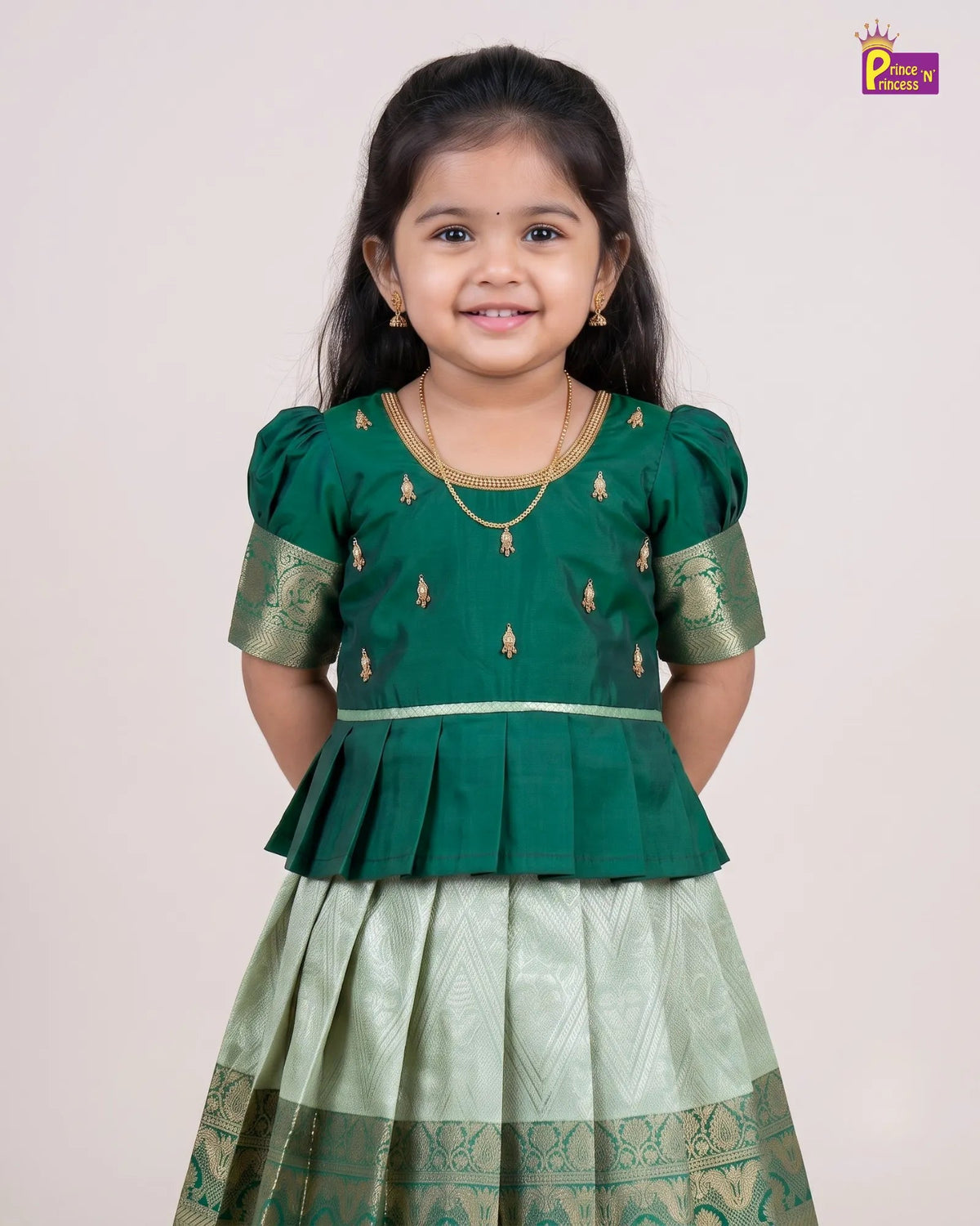 A young girl modeling a traditional Indian outfit consisting of a dark green peplum top with gold embroidery and a light green skirt with gold borders, set against a plain light beige background.