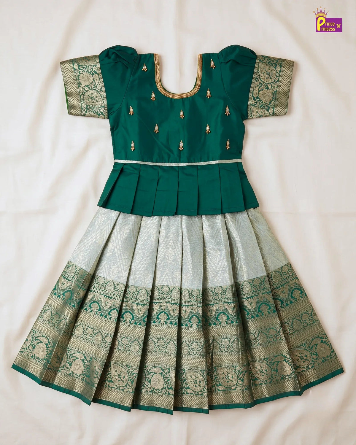 A traditional Indian girl's dress (Anarkali) featuring a dark teal green peplum bodice with gold embroidery and a pleated light silver-gray skirt with a wide teal and gold patterned border, laid flat on a white fabric background.