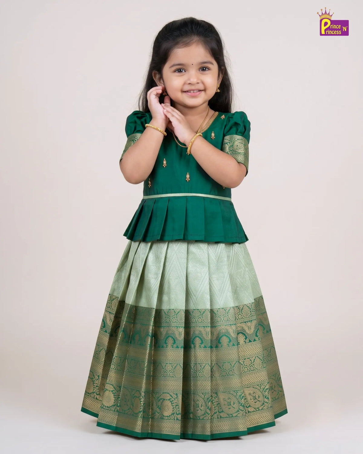 A young girl modeling a traditional Indian outfit consisting of a dark green peplum top and a light green flared skirt with gold zari patterns, set against a plain beige studio background.