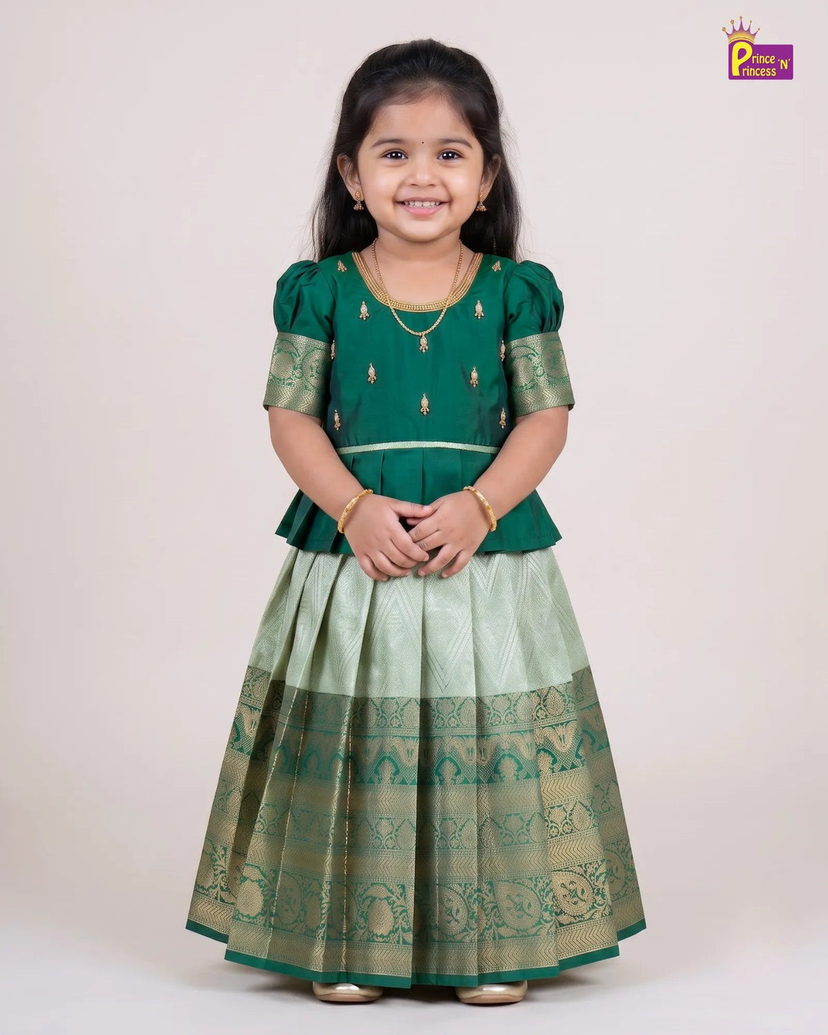 A young girl modeling a traditional Indian ethnic dress consisting of a dark green peplum top with gold embroidery and a light green pleated skirt with a wide green and gold patterned border, set against a plain beige studio background.