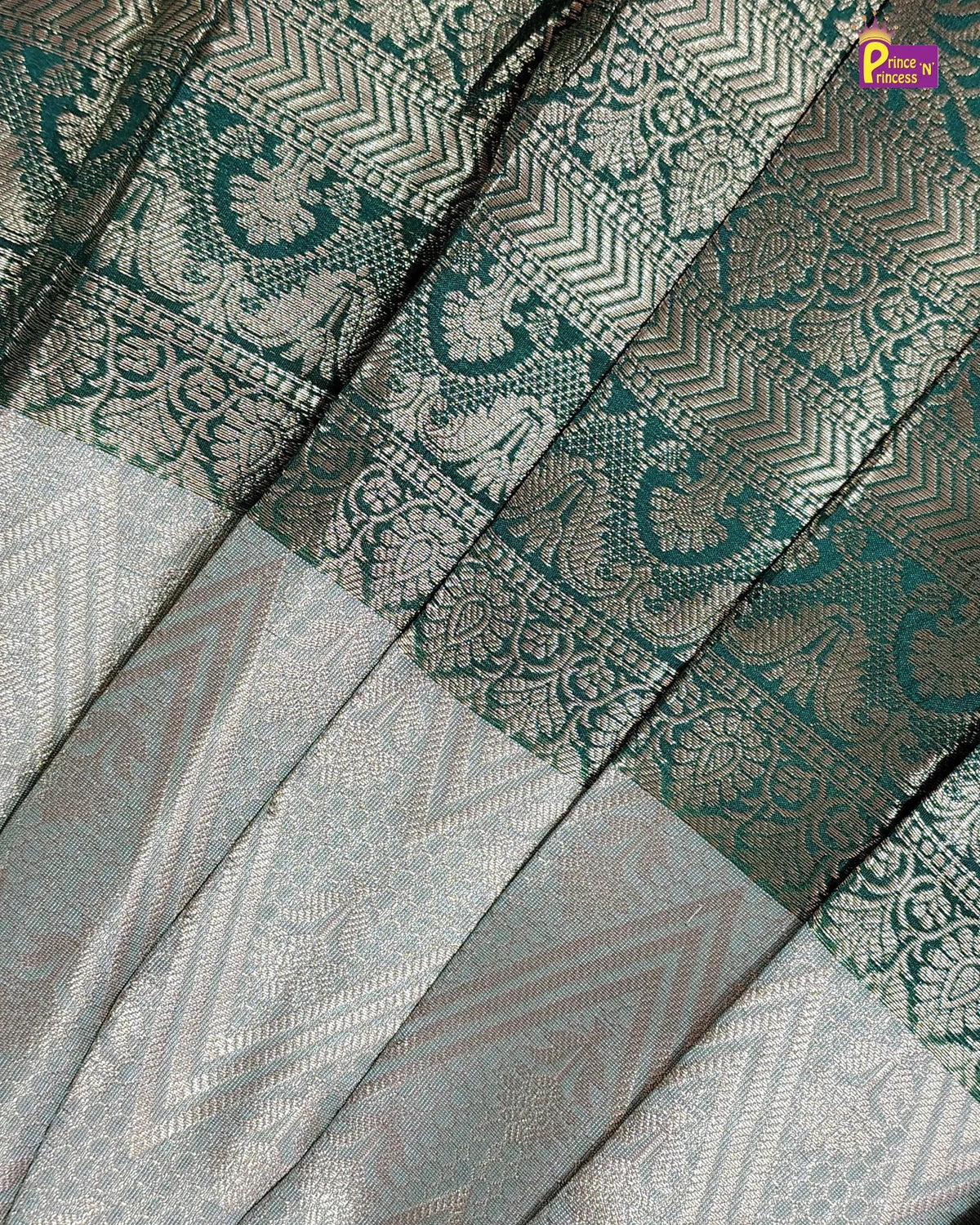 A close-up of a woven textile with a teal and silver paisley border and a light silver-gray patterned body, shown in pleated folds.