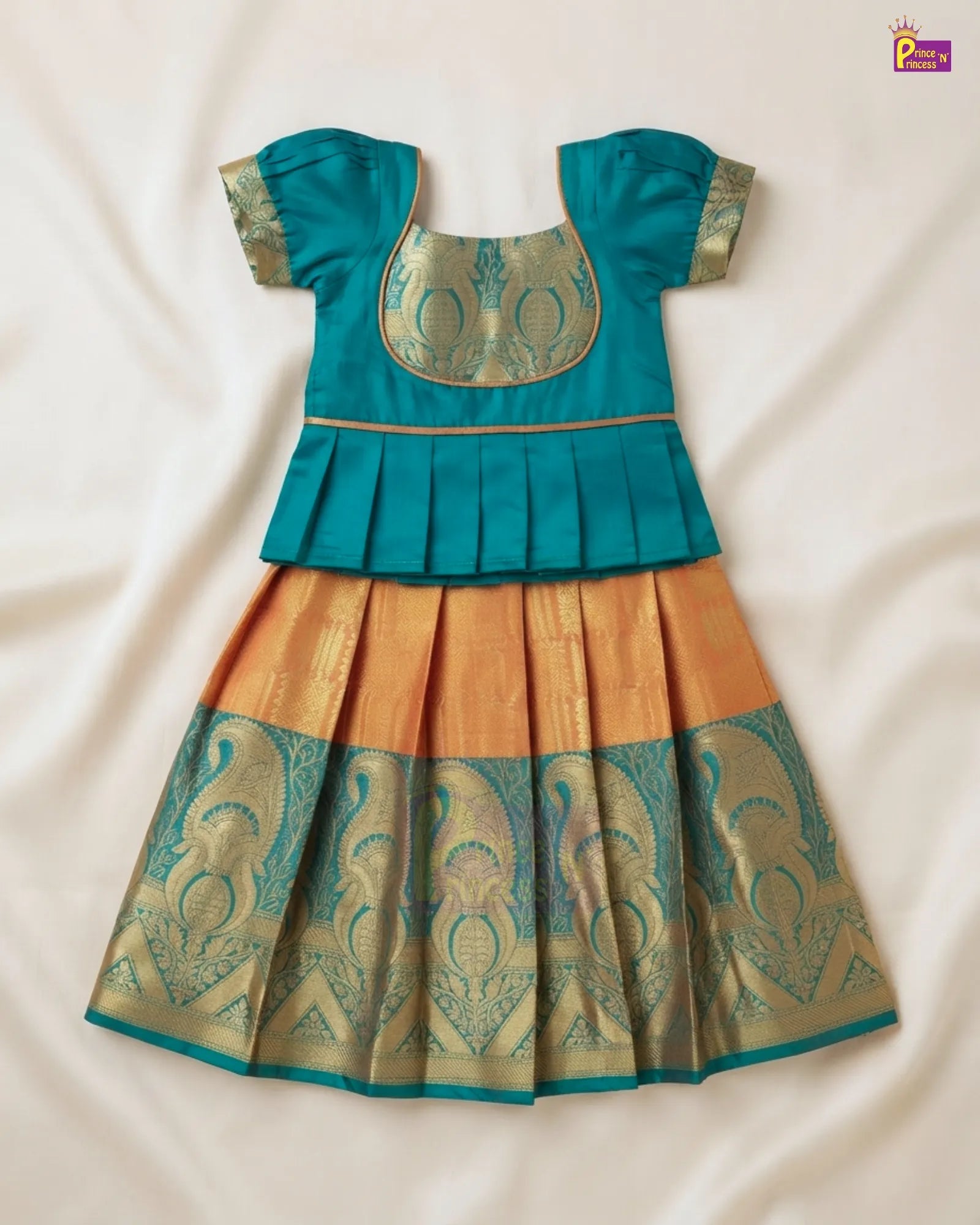 A traditional Indian girl's dress (Anarkali) laid flat on a cream-colored fabric background. The dress features a teal blue top with gold zari embroidery, a pleated peplum waist, and a flared skirt in gold and teal with intricate woven patterns.