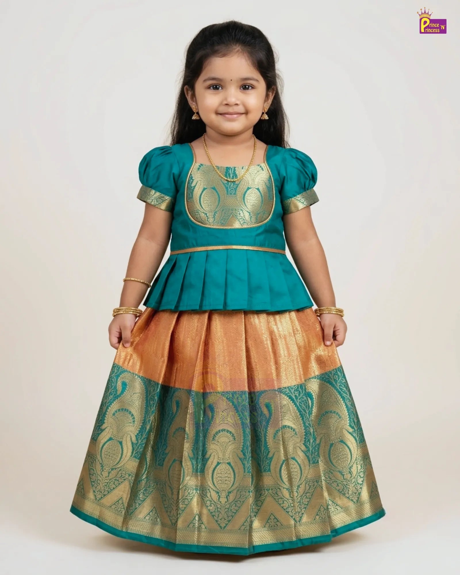 A young girl modeling a traditional Indian ethnic outfit consisting of a teal blue peplum top with gold embroidery and a matching flared skirt in gold and teal with woven patterns, set against a plain light background.