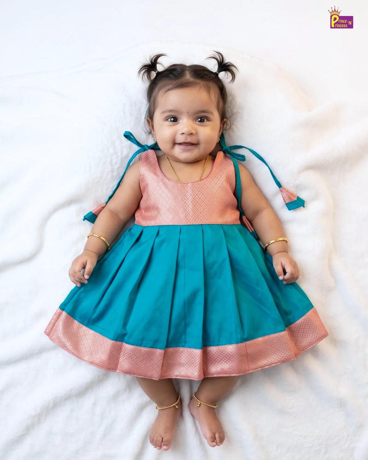 A baby girl wearing a sleeveless teal and peach kurta dress with gold bangles and a gold necklace, lying on a white blanket.