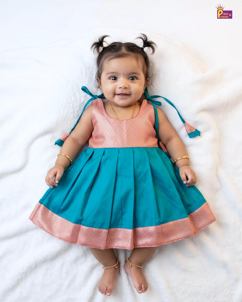 A baby girl wearing a sleeveless teal and peach kurta dress with gold bangles and a gold necklace, lying on a white blanket.