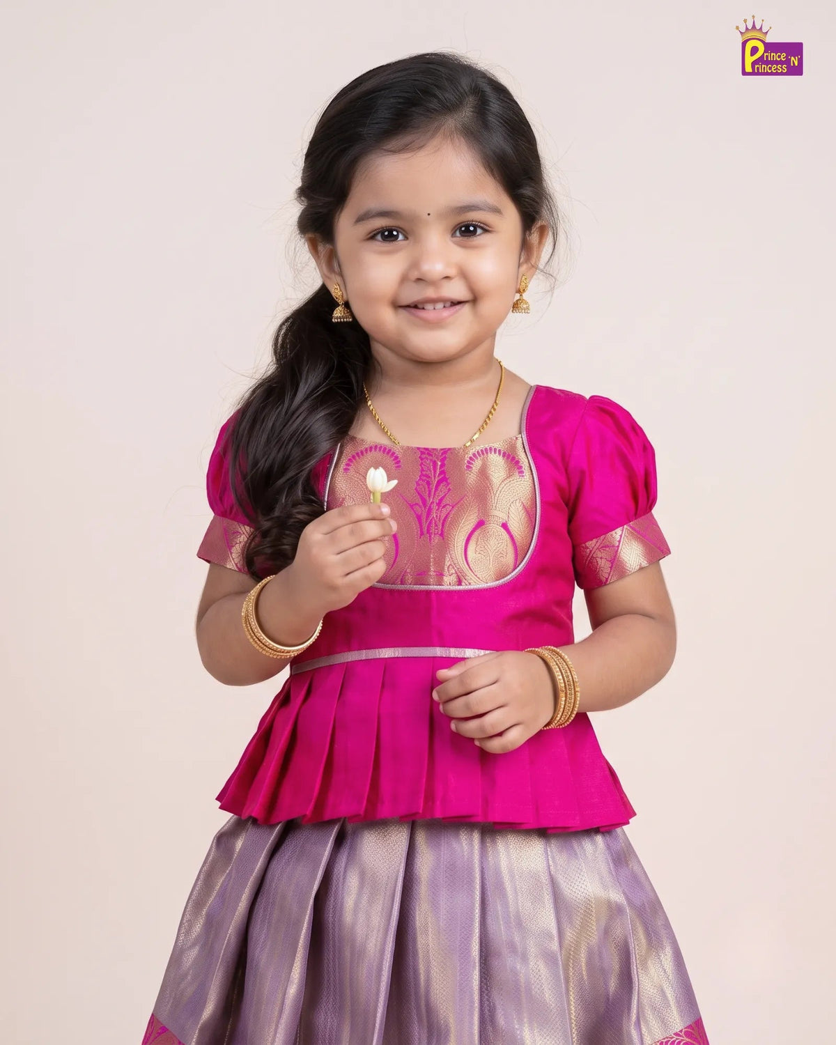 A young girl modeling a traditional Indian outfit consisting of a bright pink peplum top with gold embroidery and a matching purple skirt, set against a plain light beige background.