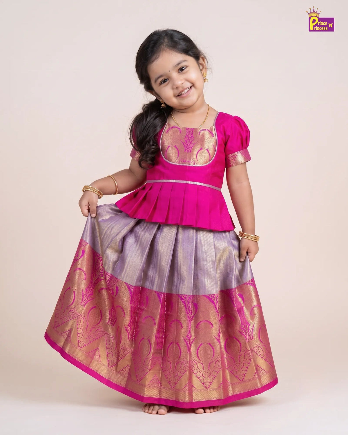 A young girl modeling a traditional Indian ethnic wear set consisting of a bright pink peplum-style blouse and a matching flared skirt with purple and gold stripes and metallic embroidery, set against a plain beige studio background.