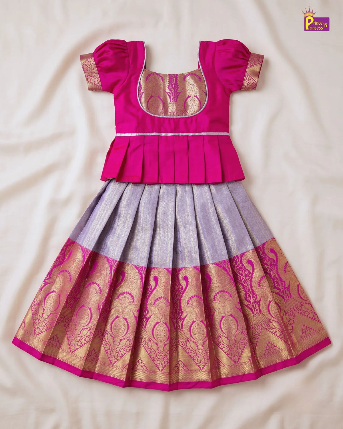 A traditional Indian girl's dress (lehenga choli) laid flat on a cream-colored fabric background. The top portion is a vibrant magenta pink with gold zari embroidery and a square neckline.