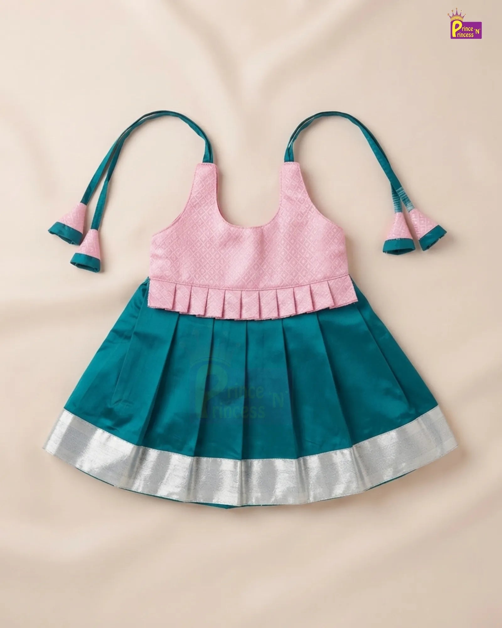 A toddler girl's sleeveless dress with a light pink textured top and a teal pleated skirt with a silver border, laid flat on a beige fabric background.