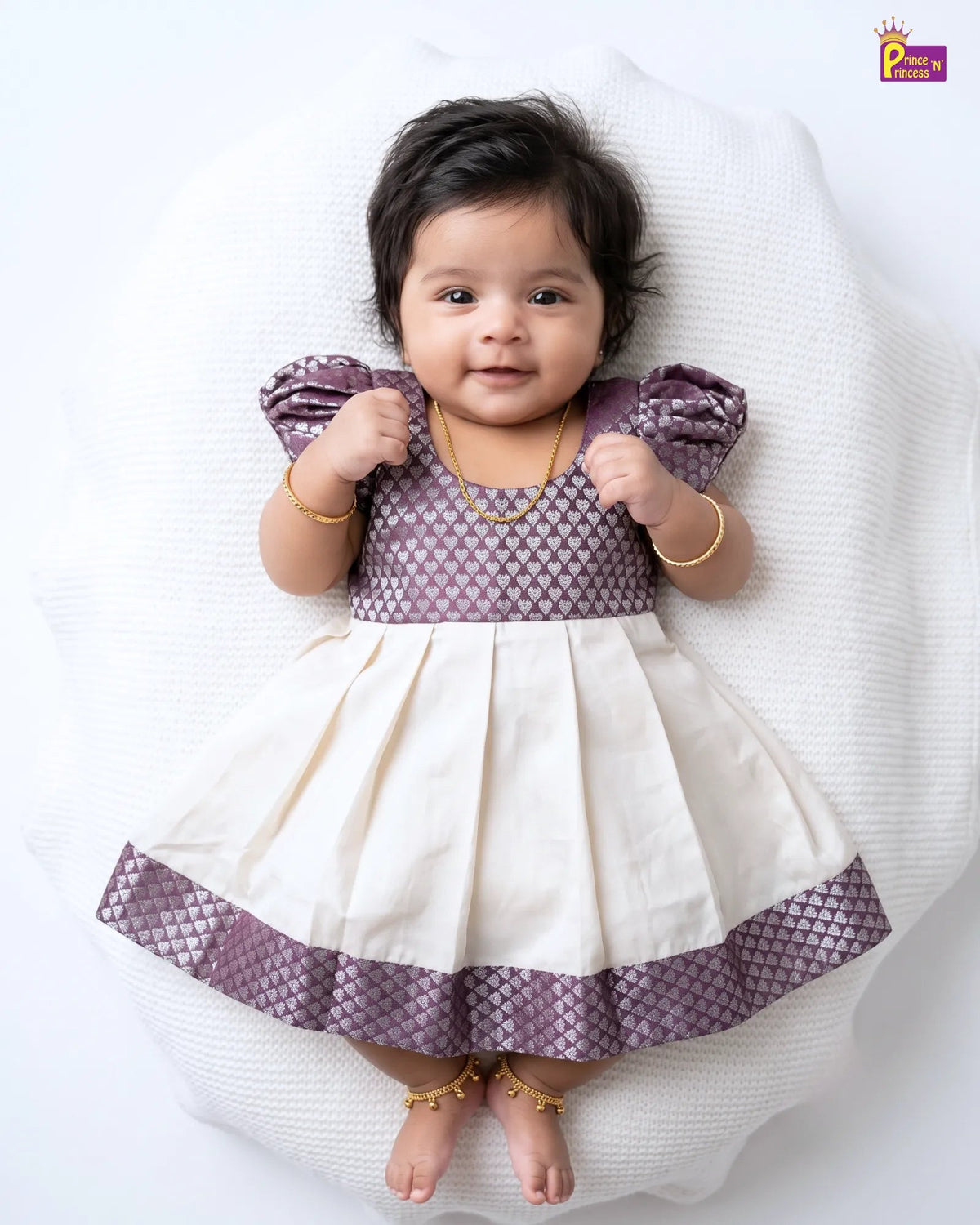 A baby girl lying on a white textured cushion, wearing a purple and cream tiered dress with gold jewelry.