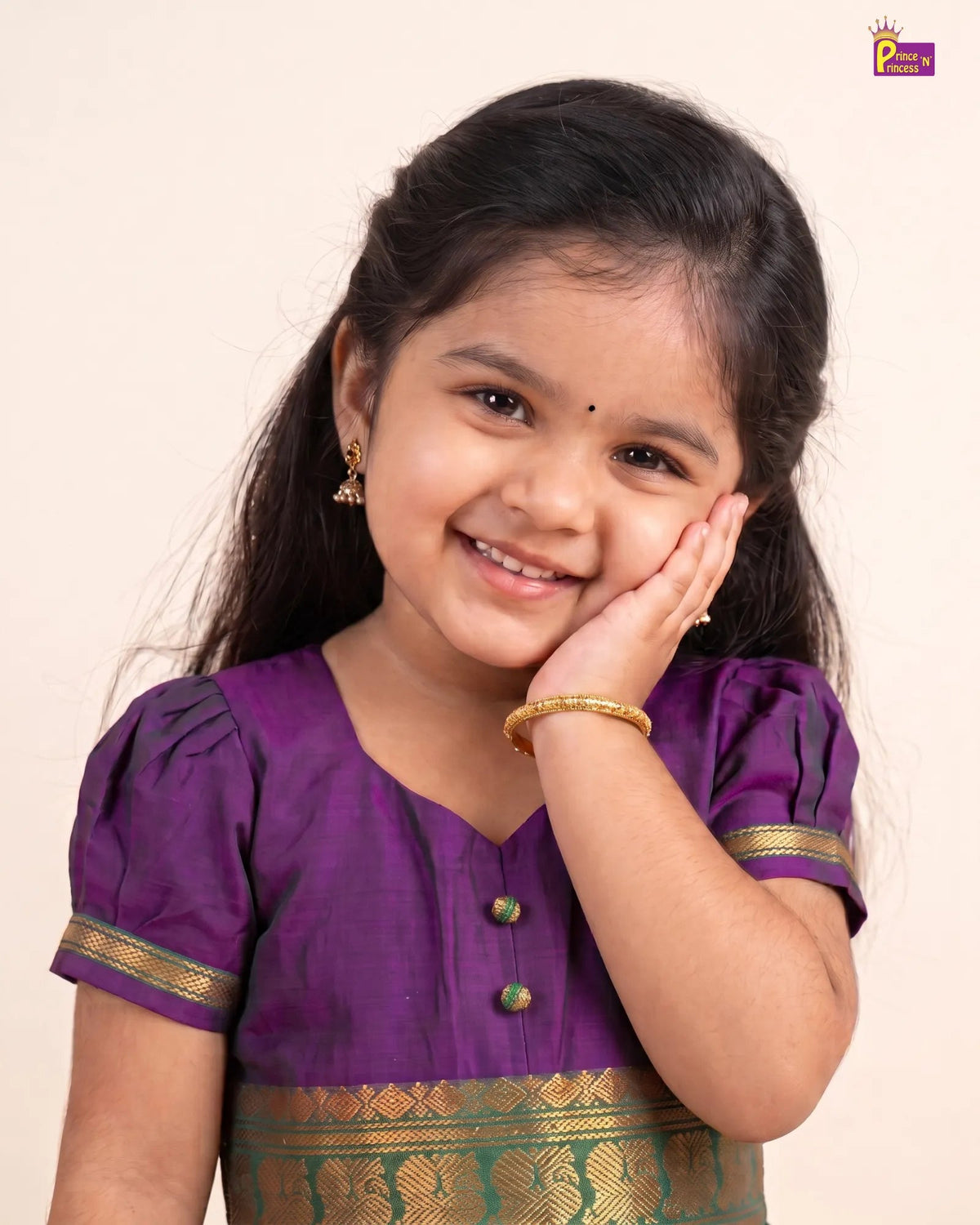 A young girl smiling and posing in a purple traditional Indian blouse with gold trim, set against a plain cream background.