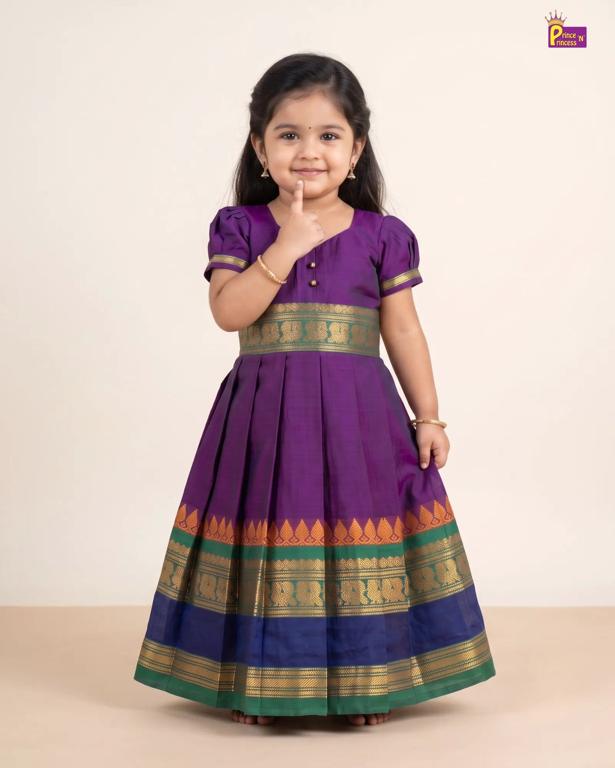 A young girl modeling a purple traditional dress with gold and green borders, standing against a plain beige studio background.